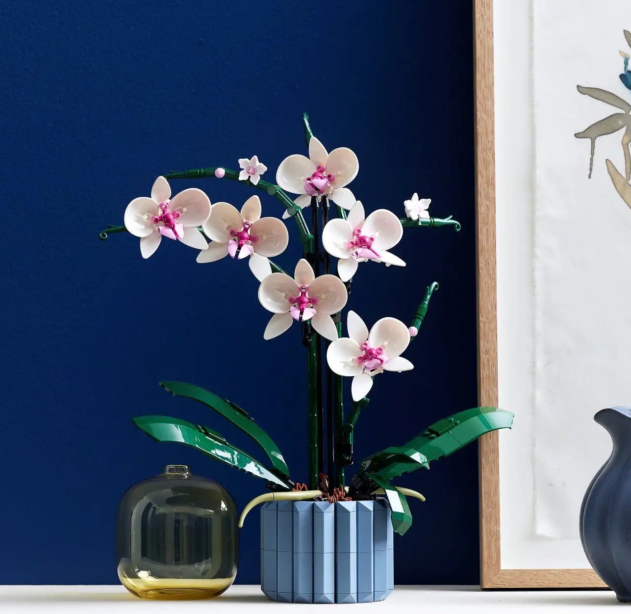 A scene with a white orchid in a pot, a flower painting, and a blue ceramic pitcher displayed on a shelf against a dark background