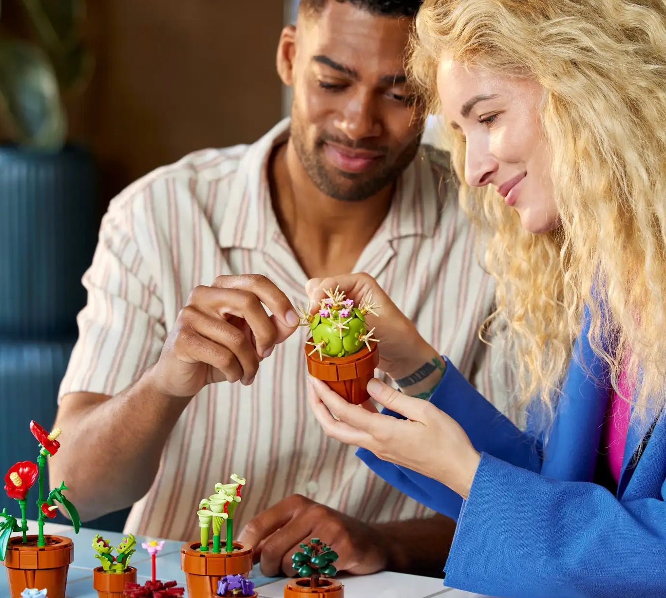 A man and a woman are sitting at a table assembling small plant-like LEGO pieces. The man holds a piece, and the woman smiles while holding a small LEGO plant
