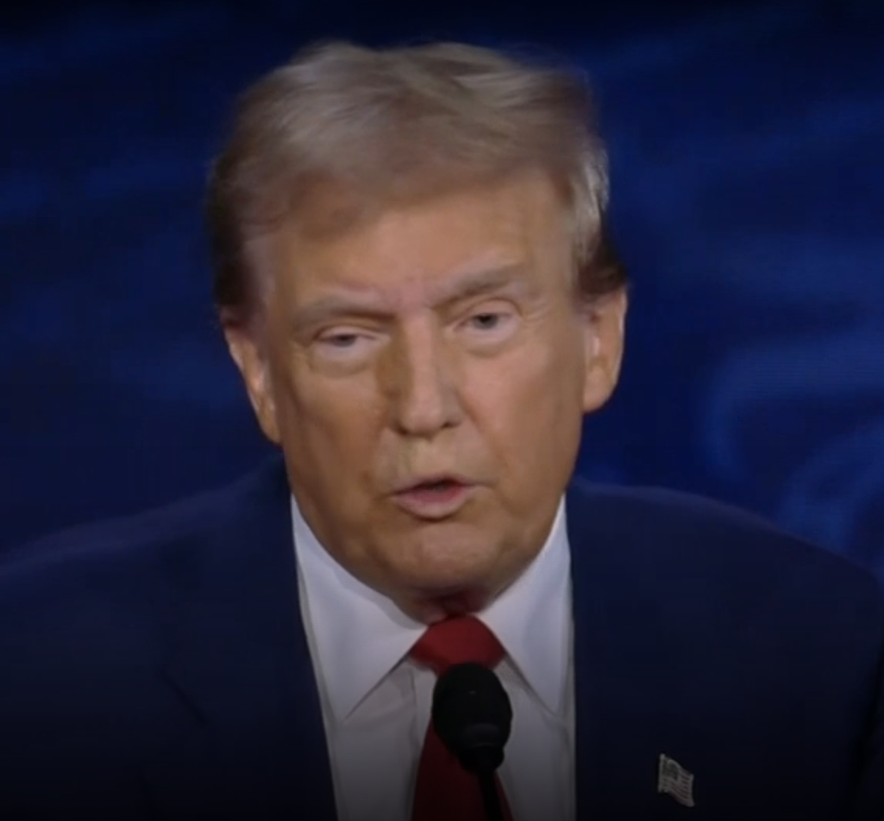 Donald Trump speaks at an event, wearing a suit and red tie