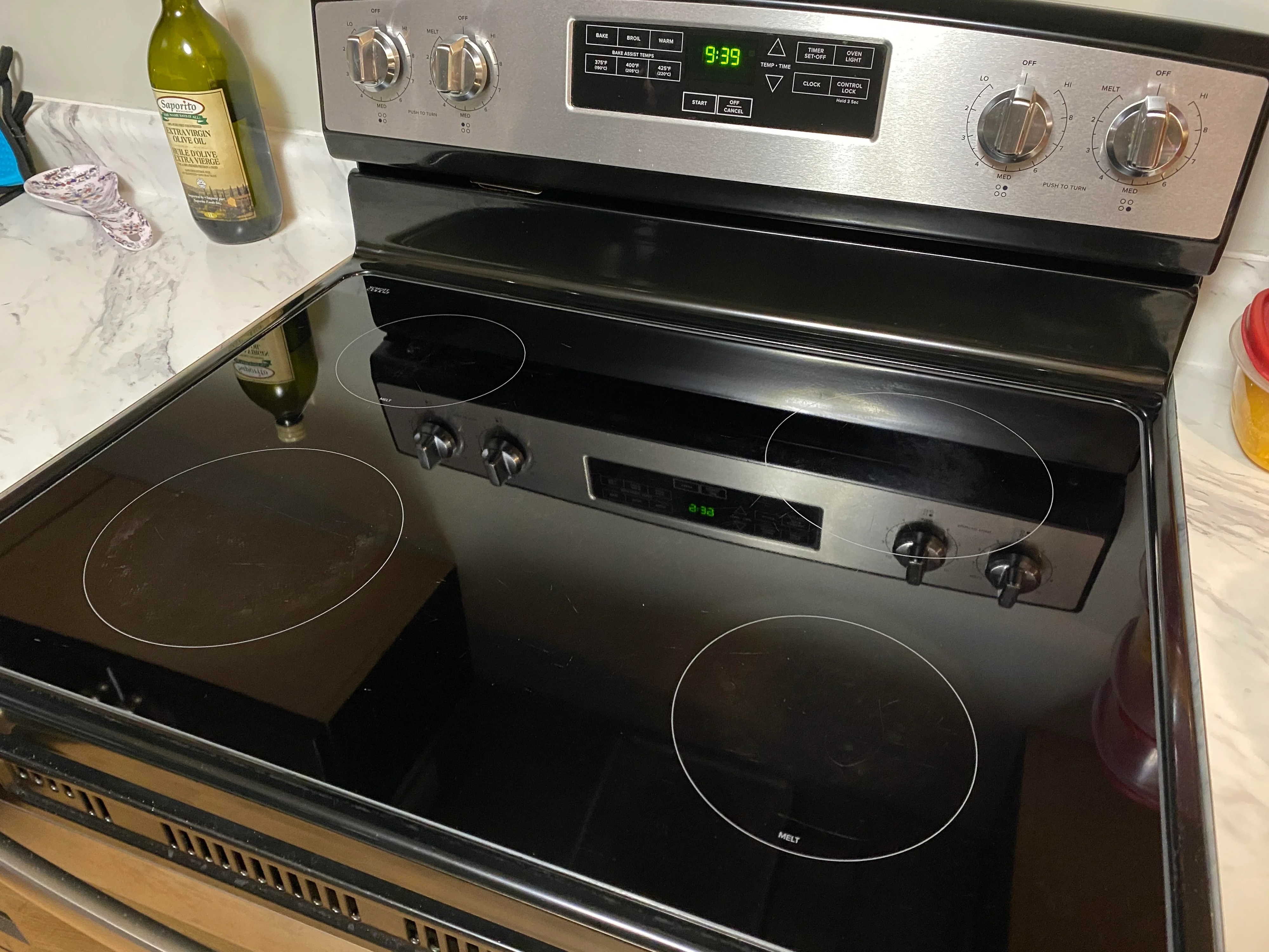 A clean stove with four burners, a digital clock displaying 3:39, and an oven below. A bottle of olive oil and other kitchen items are on the counter