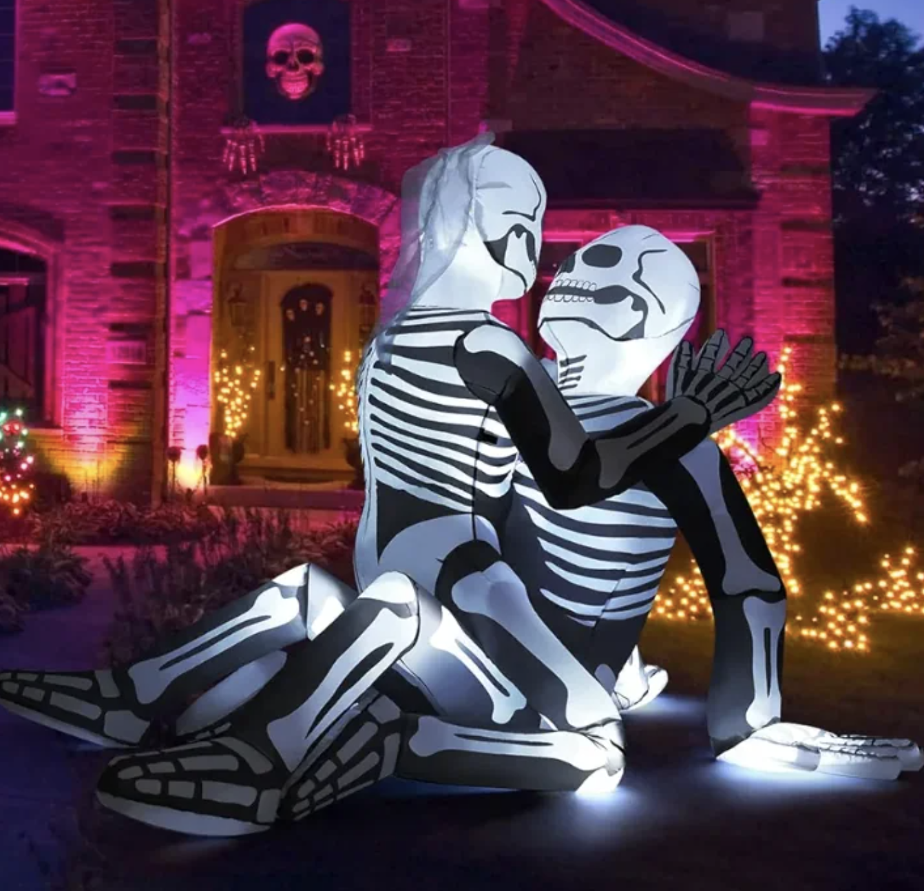 Inflatable Halloween decoration of two friendly skeletons embracing in a yard decorated with lights and a house with a skull ornament