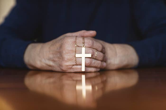 Close-up of an elderly person's hands resting on a table, holding a gold cross necklace. The person's face is not visible in the image