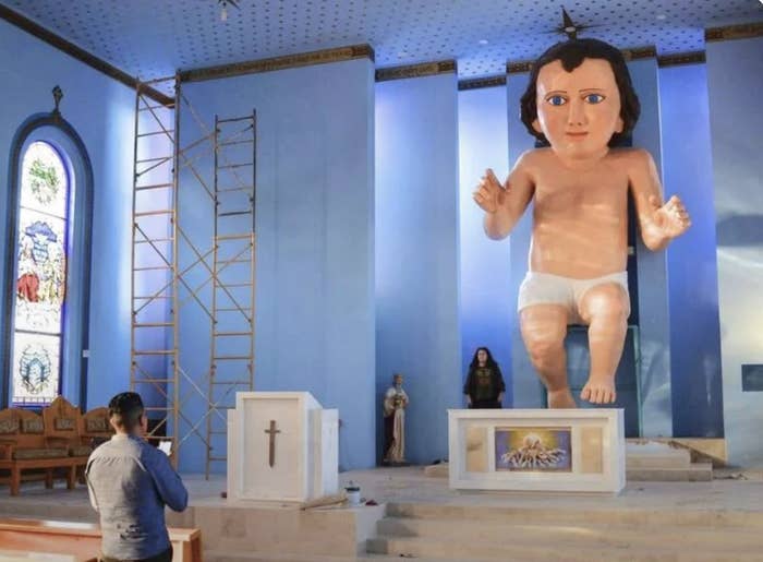 A person stands in a church with a giant baby Jesus statue in a diaper hovering above the altar. A scaffold is set up nearby