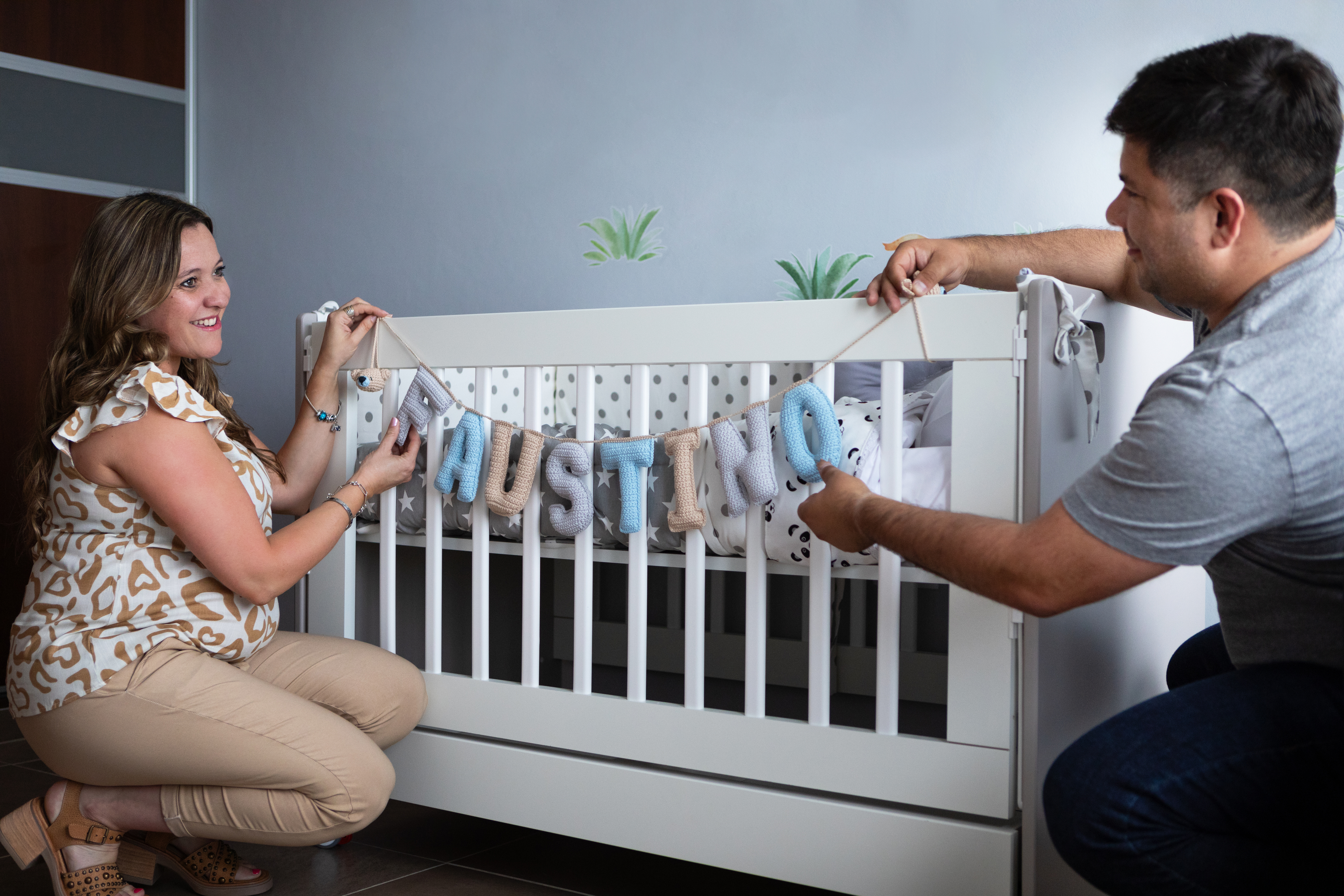 A woman and a man are decorating a white crib with a bunting that spells "Agustino."