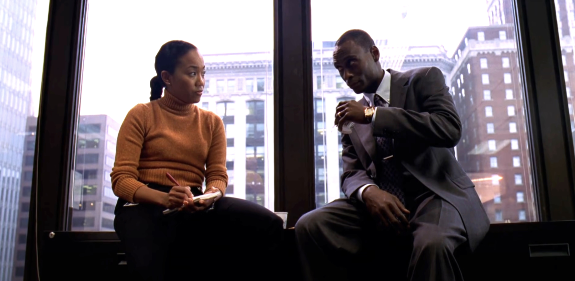 A woman, holding a notebook and pen, talks with a man in a suit by a large office window with a cityscape view in the background