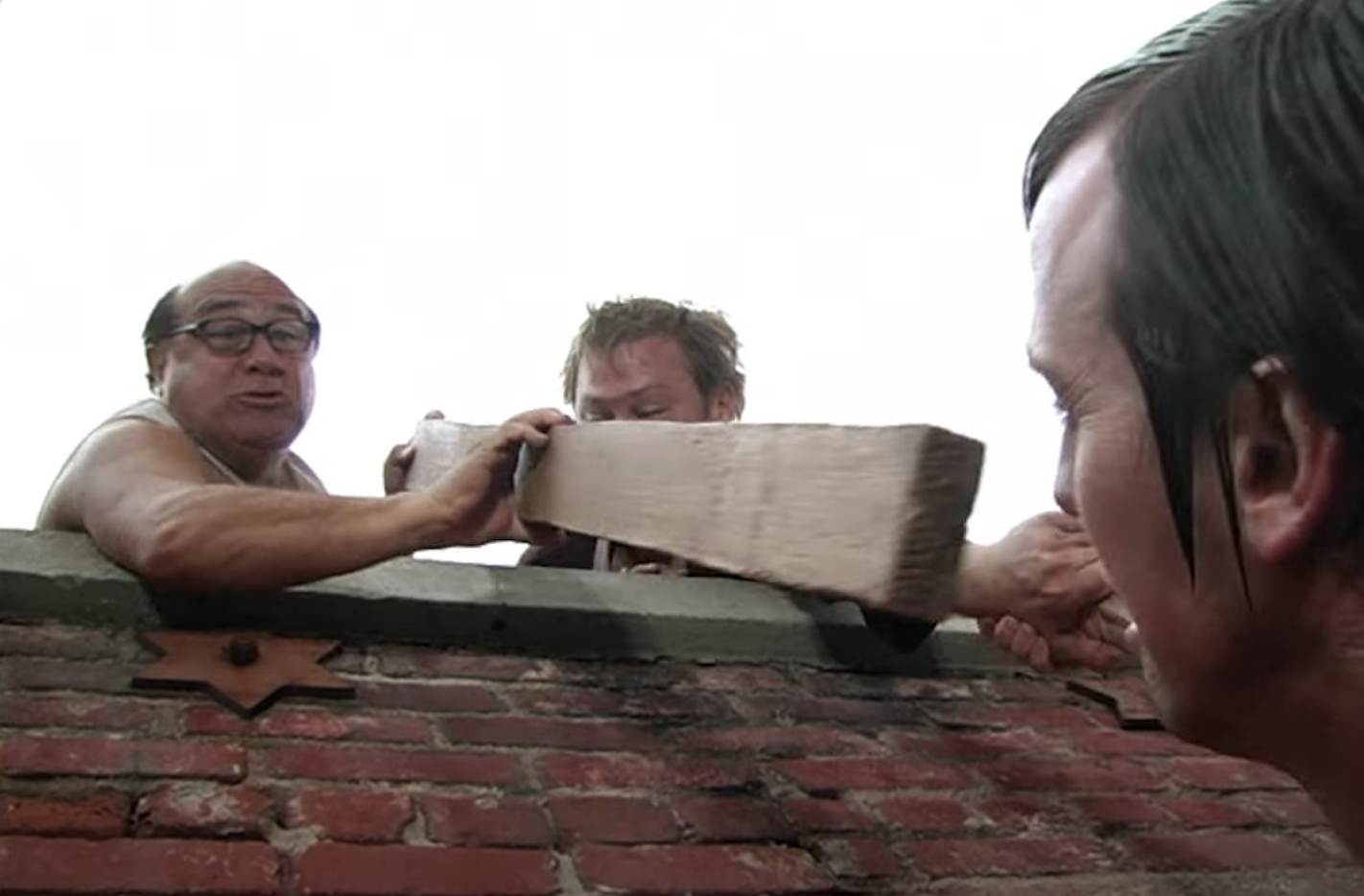 Danny DeVito and other actors in a scene from a movie or TV show, involving a wooden plank being passed over a brick wall