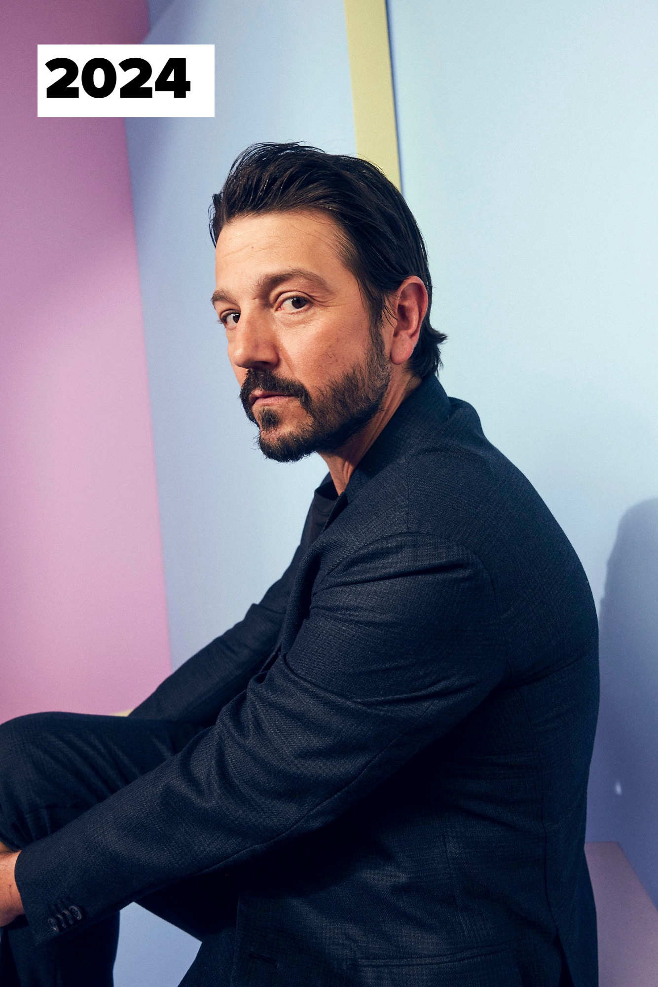 Diego Luna poses in a dark suit, sitting against a pastel-colored background