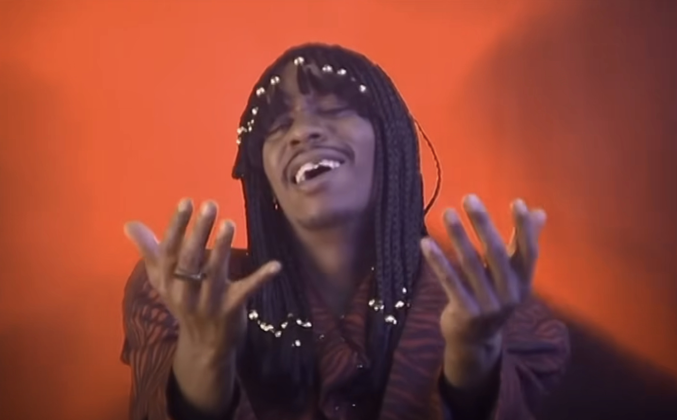 Dave Chappelle with long braided hair adorned with beads, smiling and wearing a patterned blazer, extends their hands in a gesture against a plain backdrop