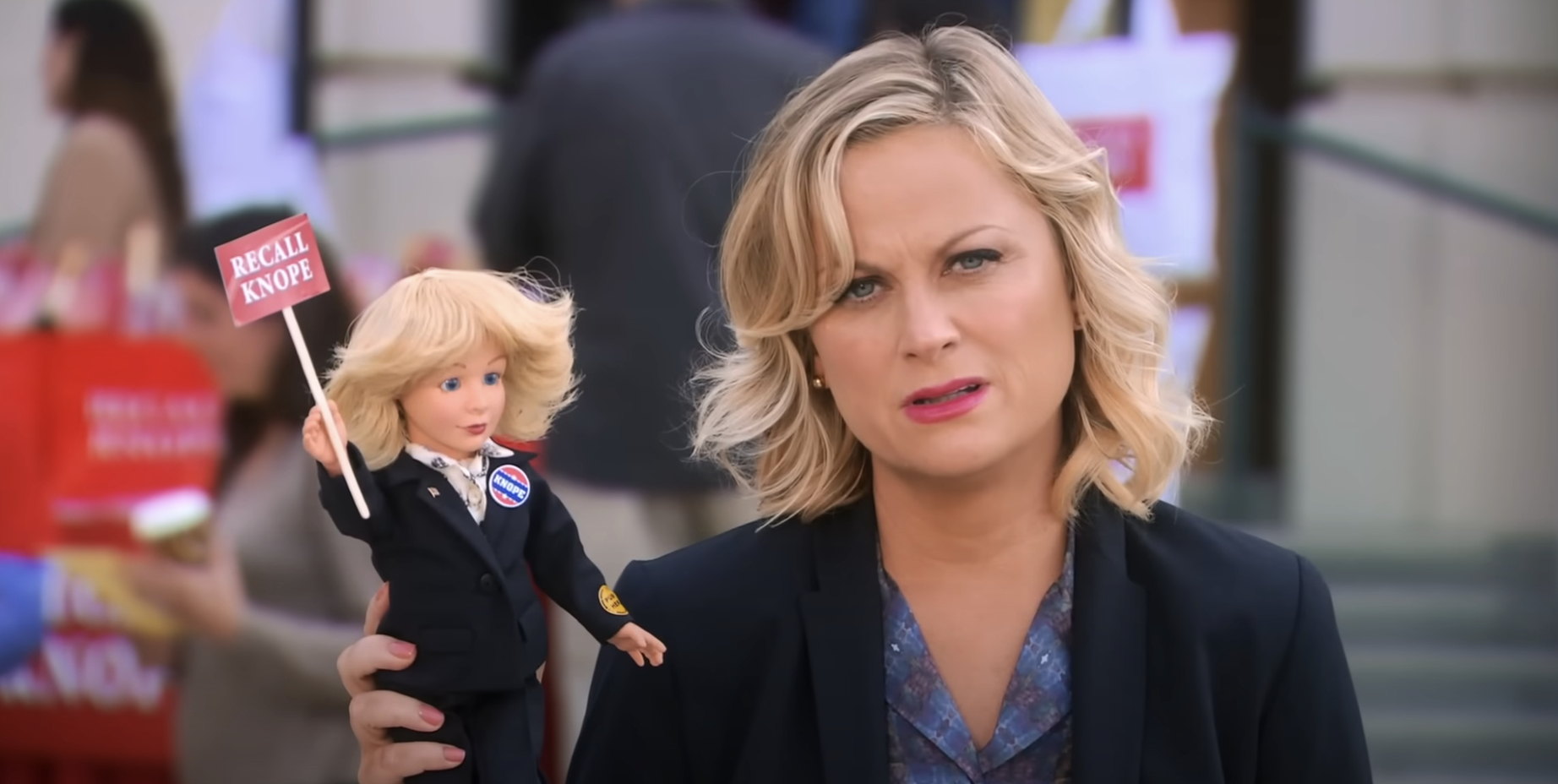 Amy Poehler, as Leslie Knope, holds a doll with a &quot;Recall Knope&quot; sign, striking a humorous pose in a scene from the show &quot;Parks and Recreation.&quot;