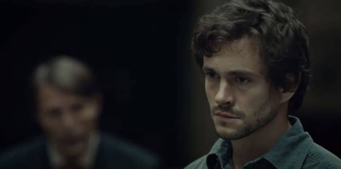Hugh Dancy, with a serious expression, in a scene from a TV show, with Hannibal out of focus, is in the background