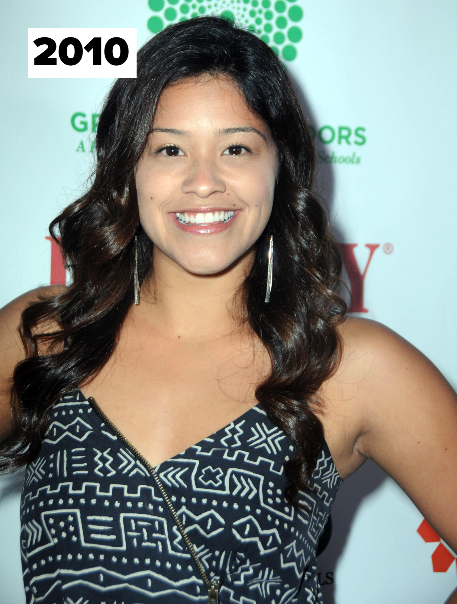 Gina Rodriguez smiles at a public event, wearing a sleeveless patterned dress and long earrings