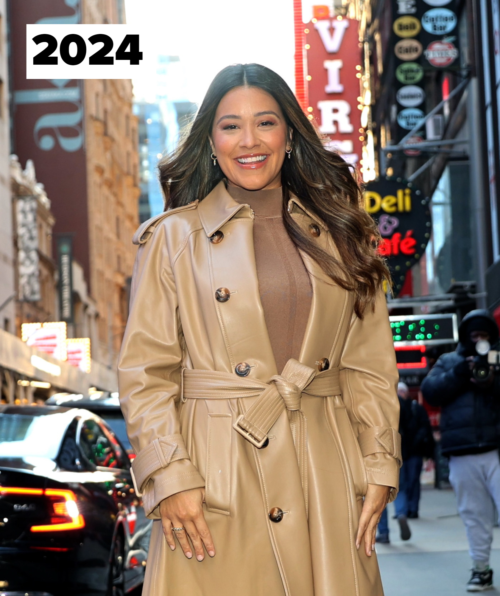 Gina Rodriguez smiles while wearing a belted trench coat over a turtleneck sweater. She stands on a city street with a bright "VIR" sign behind her