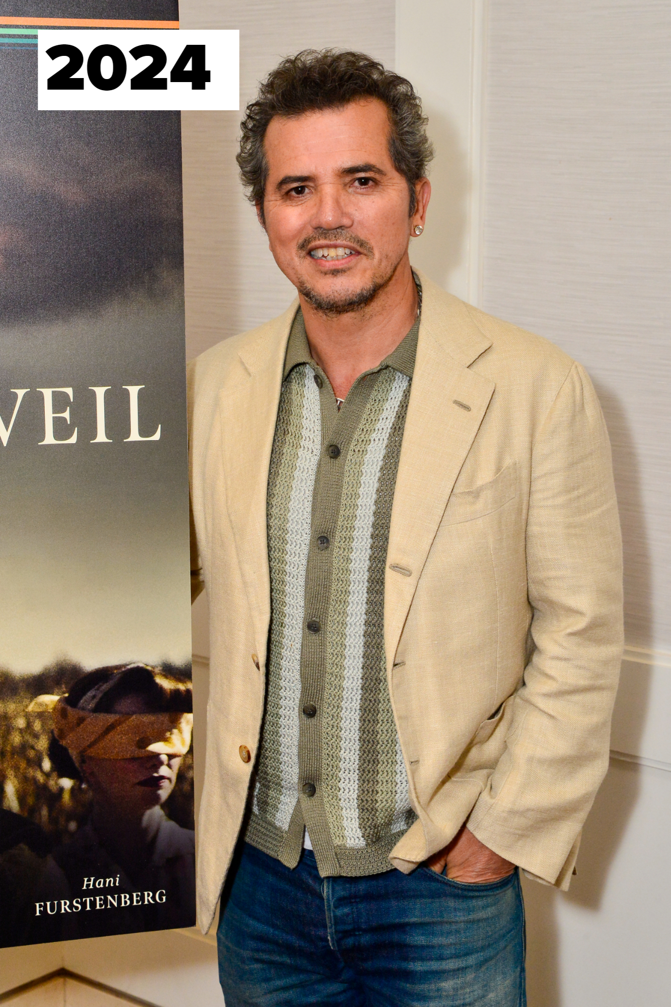 John Leguizamo stands next to a poster, wearing a beige blazer over a patterned shirt and jeans, smiling with one hand in his pocket