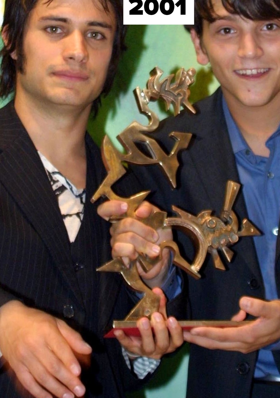 Gael Garcia Bernal and Diego Luna hold a trophy together, dressed in suits, at an event