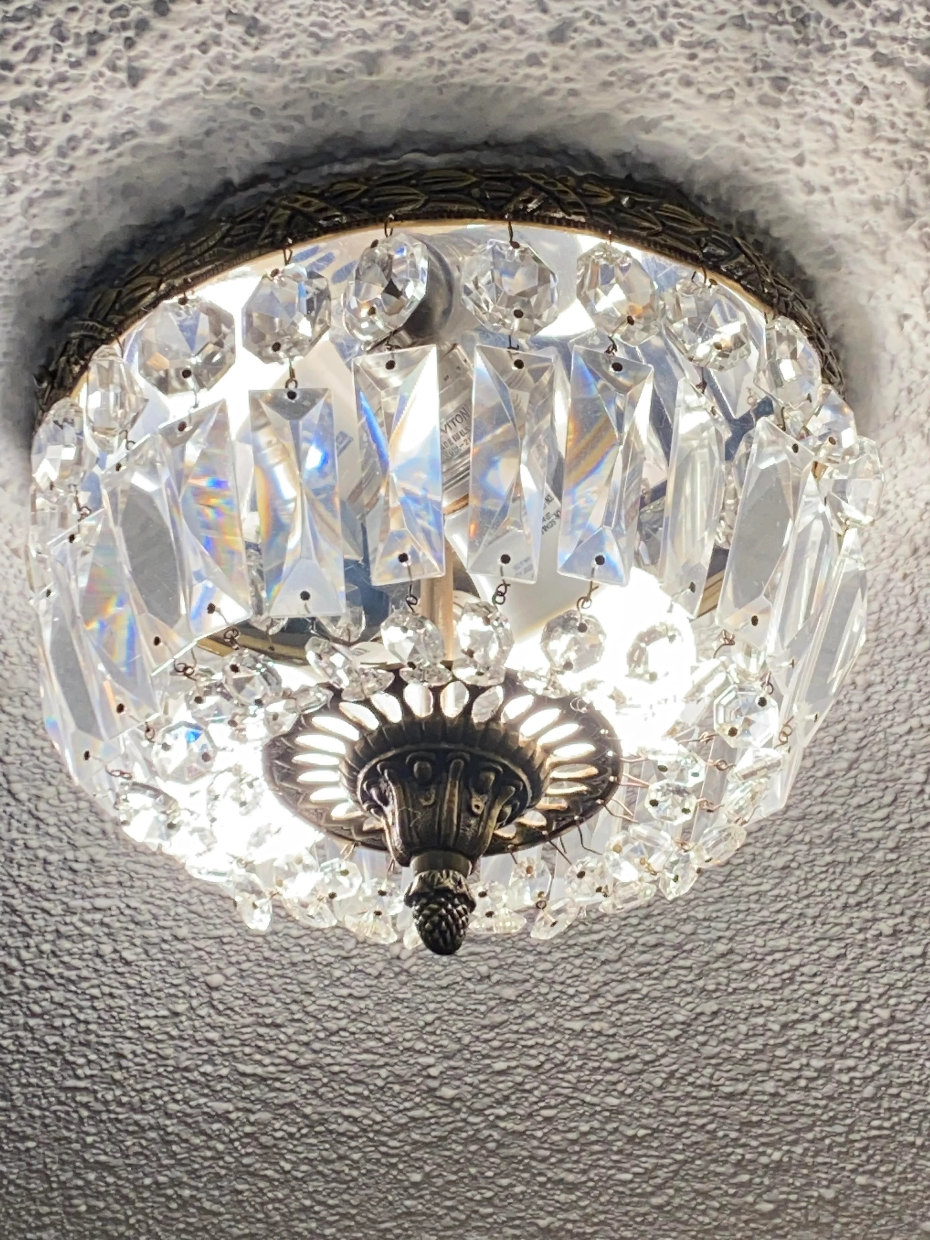 Crystal chandelier with intricate design, mounted on a textured ceiling, emitting bright light. There are no people in the image