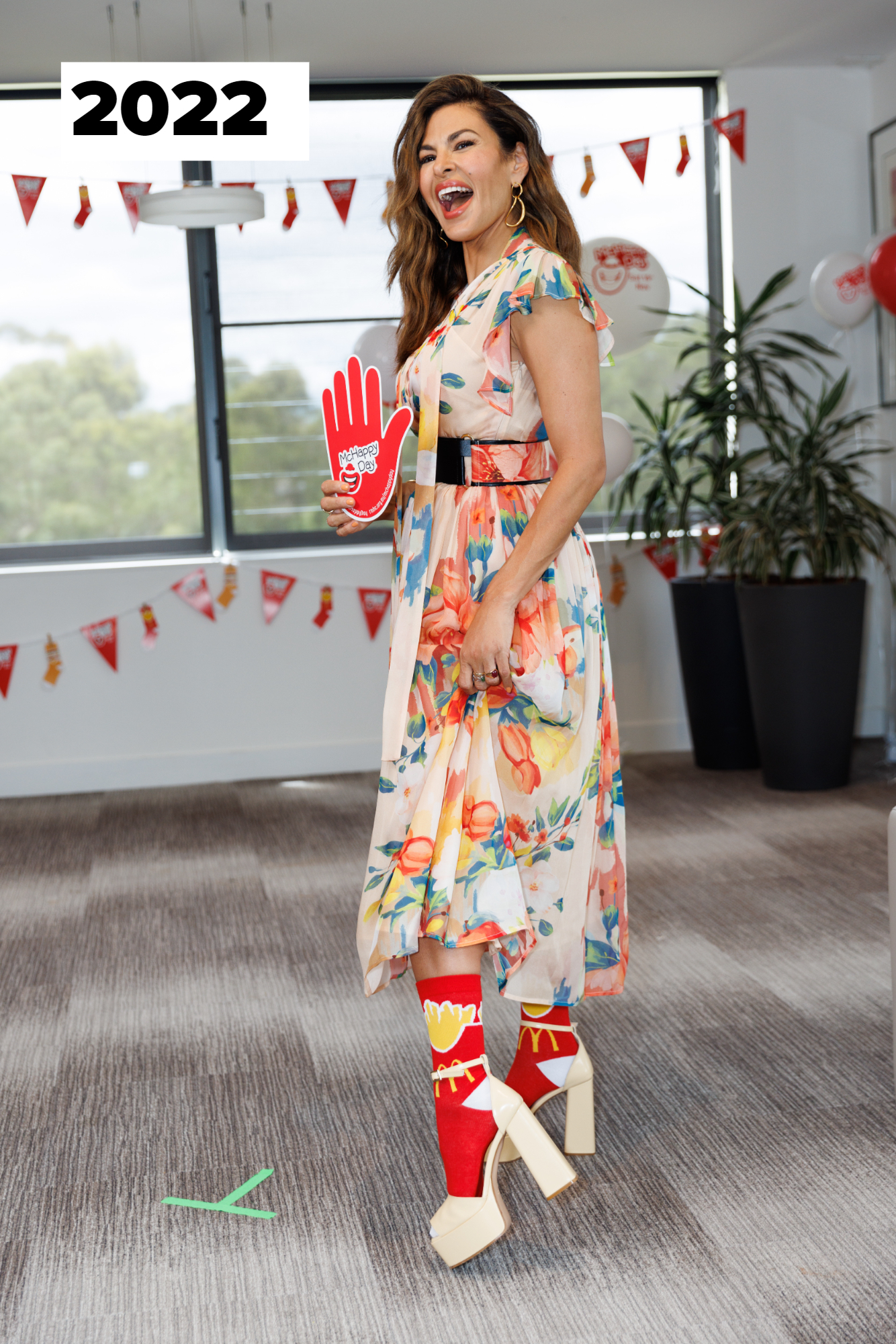 Eva Mendes, in a floral dress, holds a red foam hand and wears novelty socks with high heels. She is standing in a decorated room