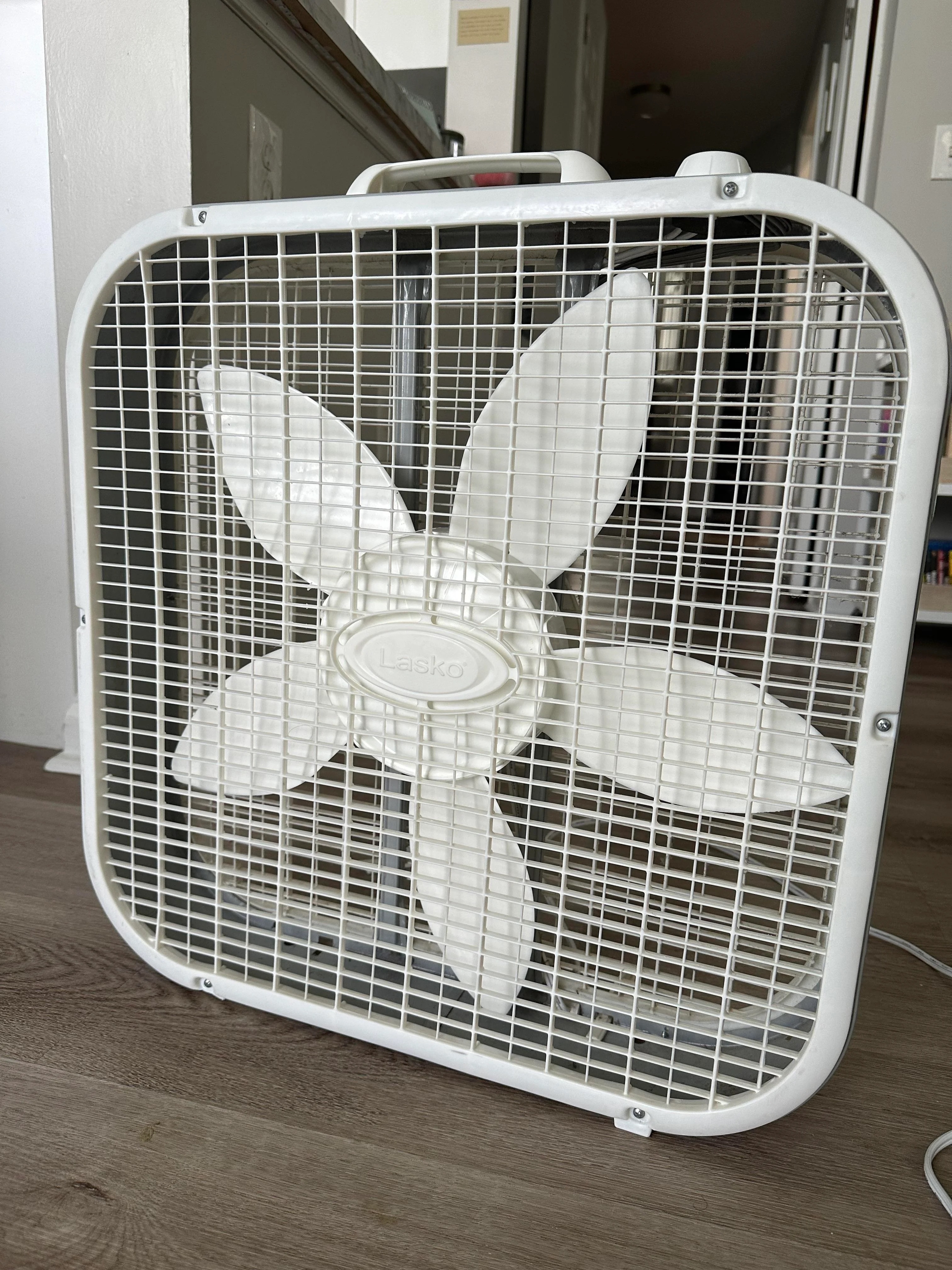 Square white Lasko box fan with flower-shaped blades placed on a wooden floor in an indoor setting