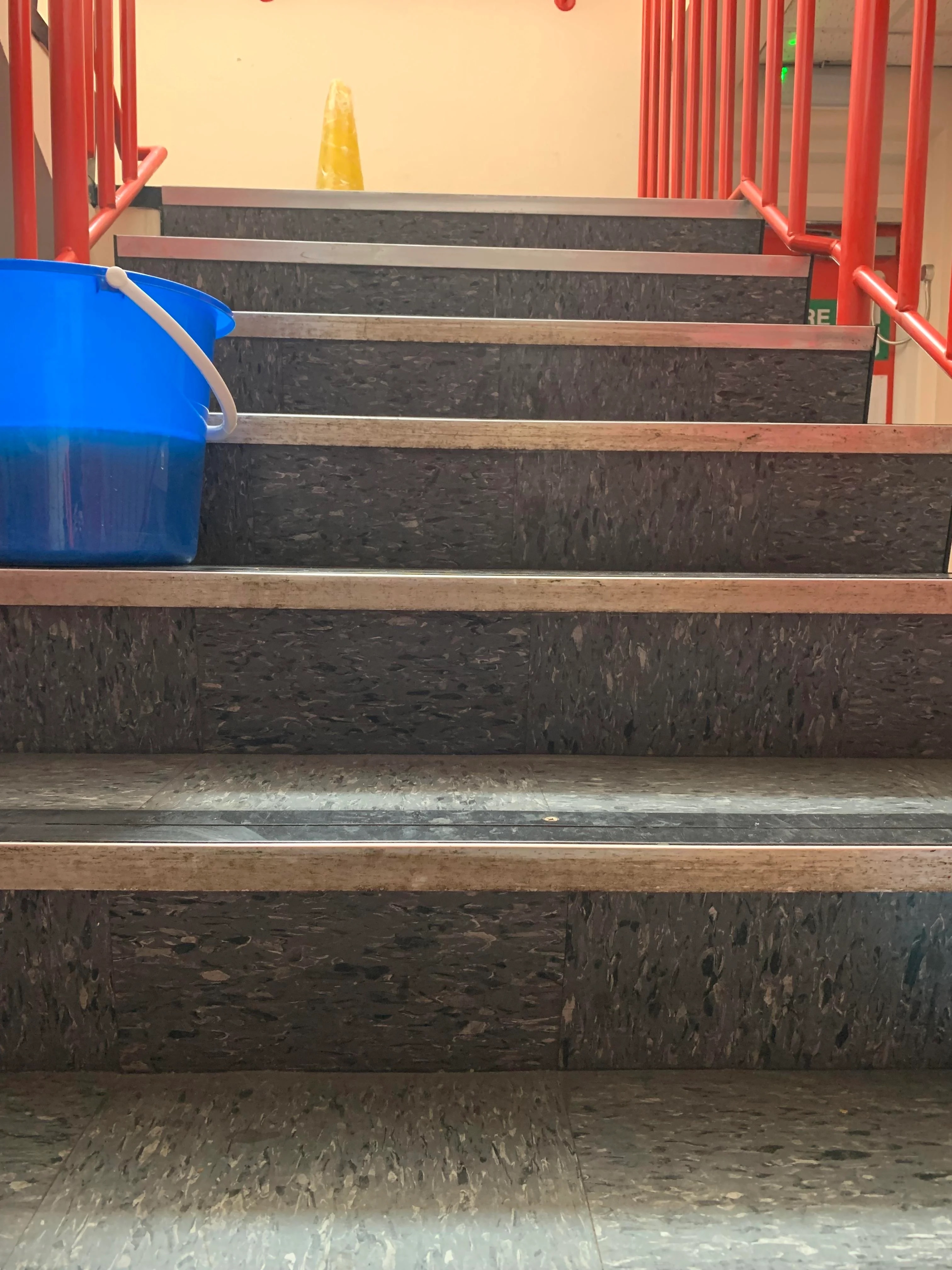 Staircase in a building with a blue bucket placed on one of the steps and red railings on the sides