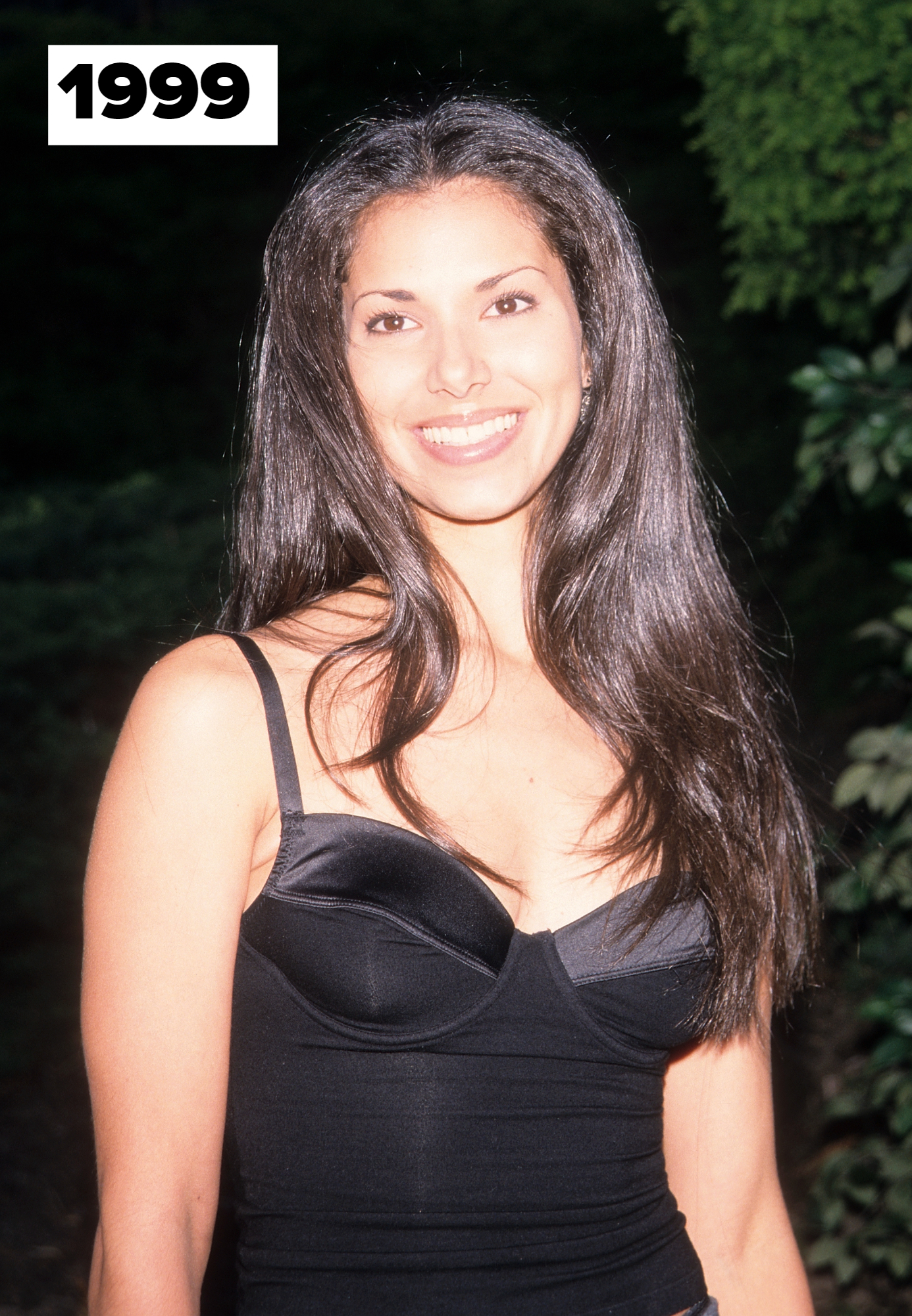 Roselyn Sanchez with long, straight hair smiles, wearing a fitted black sleeveless top. The background is foliage. Names are not provided