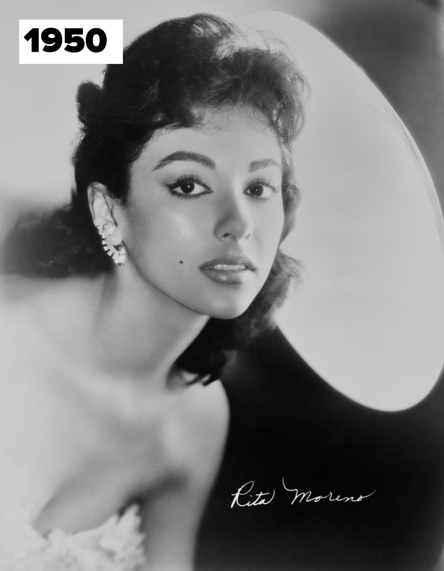Rita Moreno poses elegantly in a black and white portrait, wearing a strapless dress and diamond earrings. Her name is signed on the bottom right
