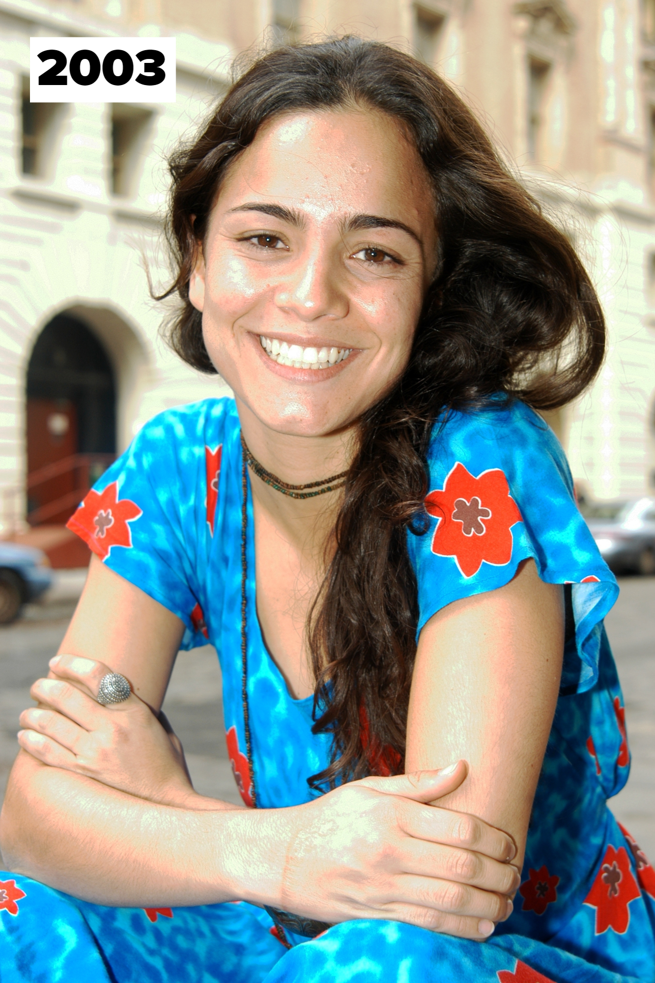 Alice Braga smiles, wearing a blue dress with red flower prints. She is seated with her arms folded, outside a building