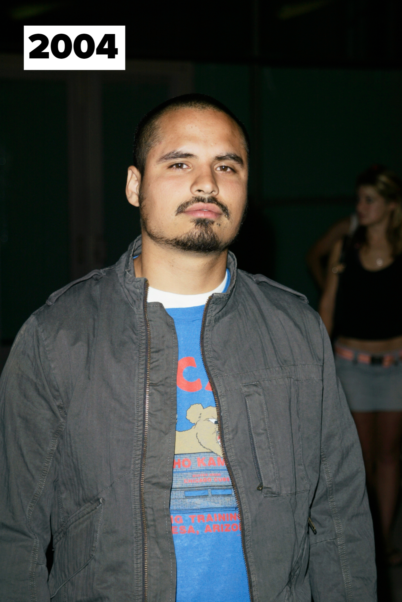 Michael Peña stands outdoors, wearing a dark jacket over a graphic t-shirt. A person in casual attire is blurred in the background