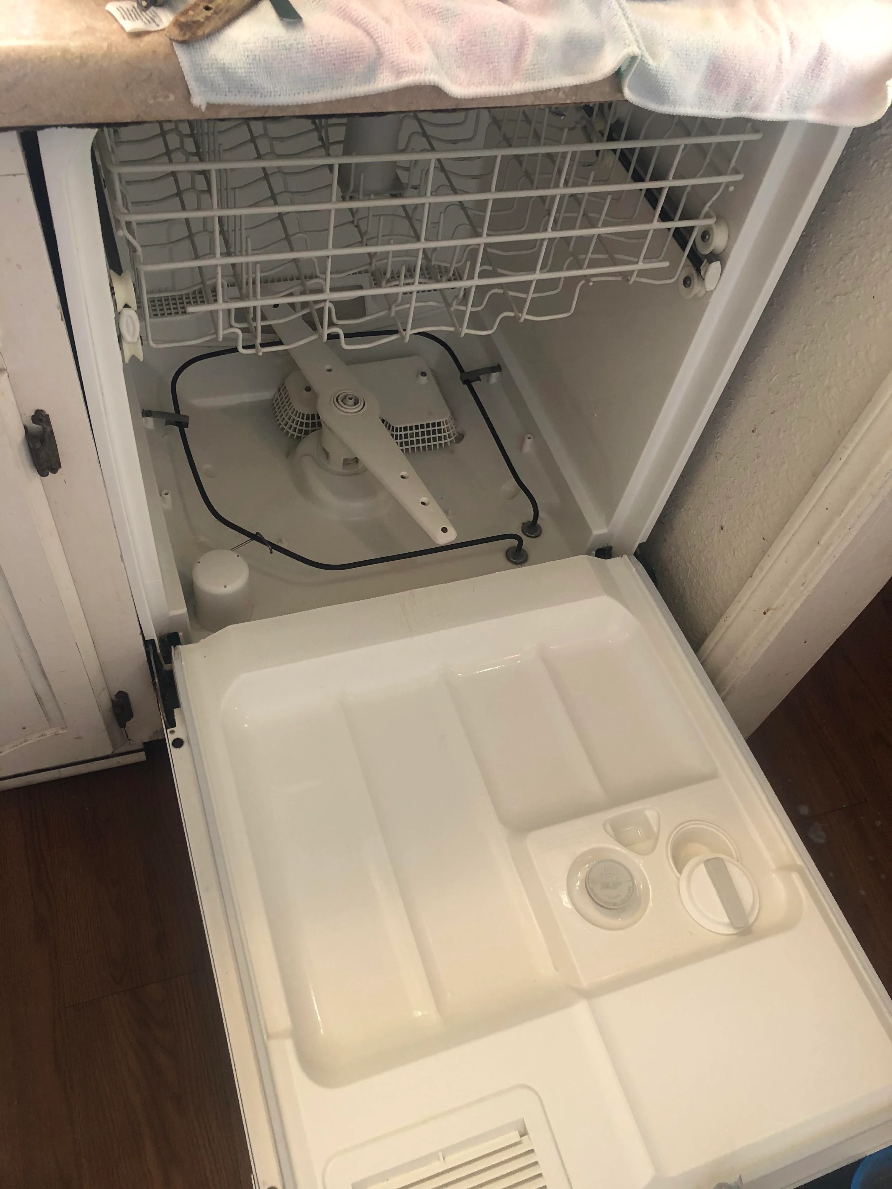 An open dishwasher with the bottom rack removed, showing the interior mechanisms. The kitchen counter and adjacent cabinets are partially visible. No people are in the image