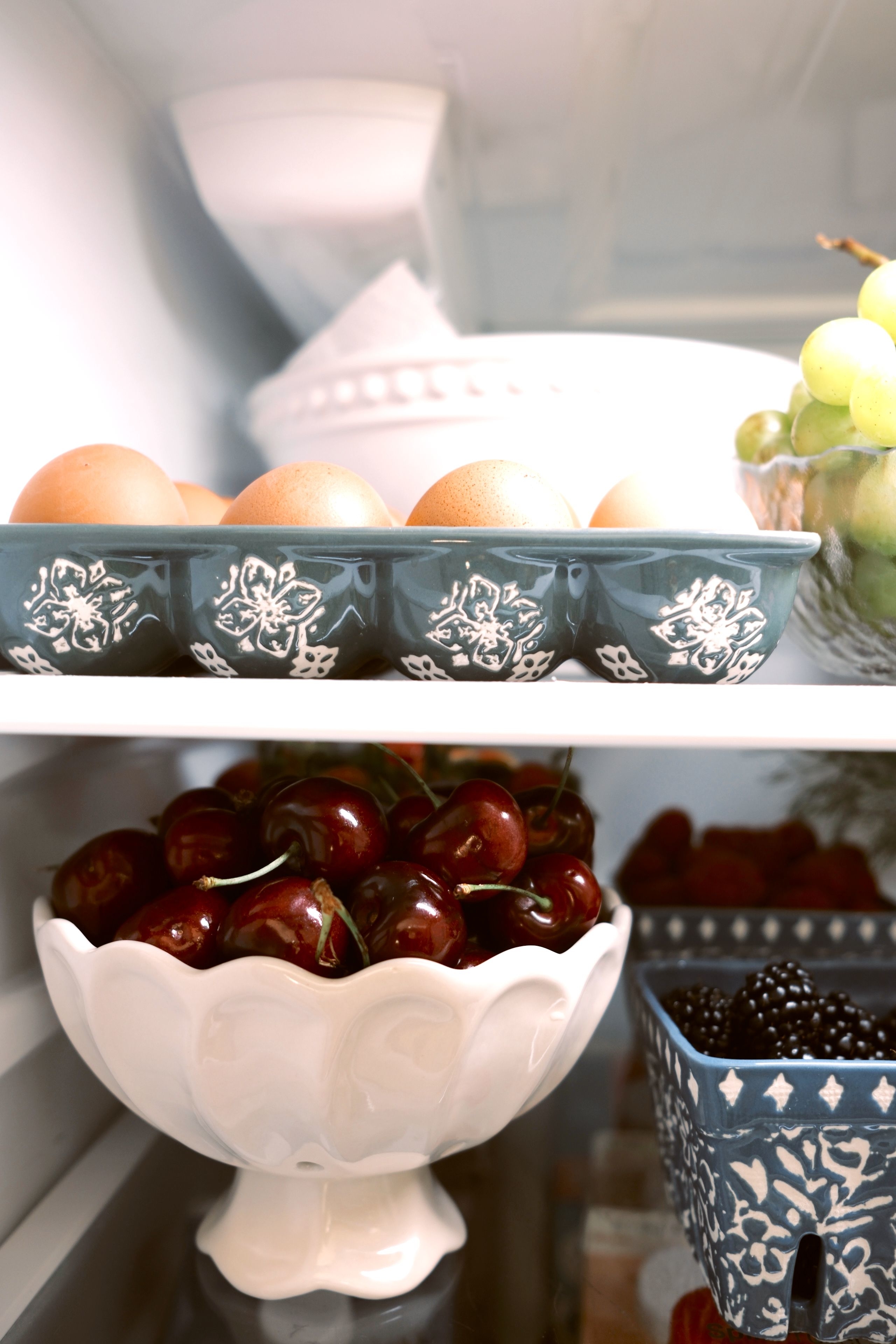 A refrigerator shelf showing ceramic containers with eggs, cherries in a white bowl, grapes, and blackberries