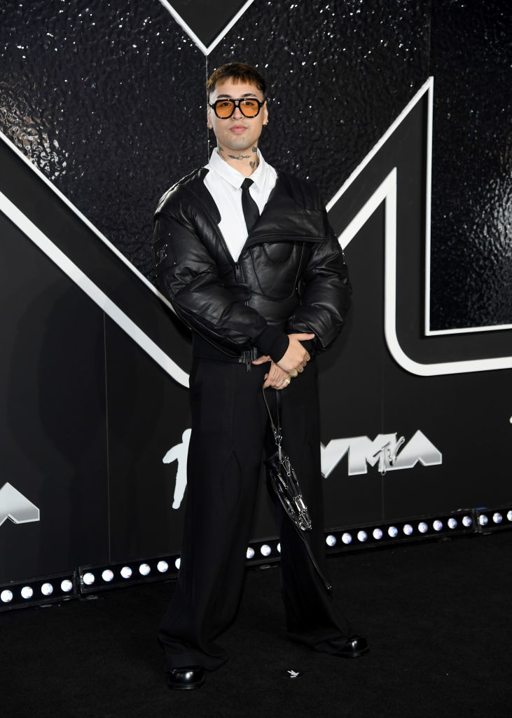 Tiago PZK on the red carpet at the VMAs, wearing wide-leg pants, a shirt, tie, and an oversized jacket with tinted glasses