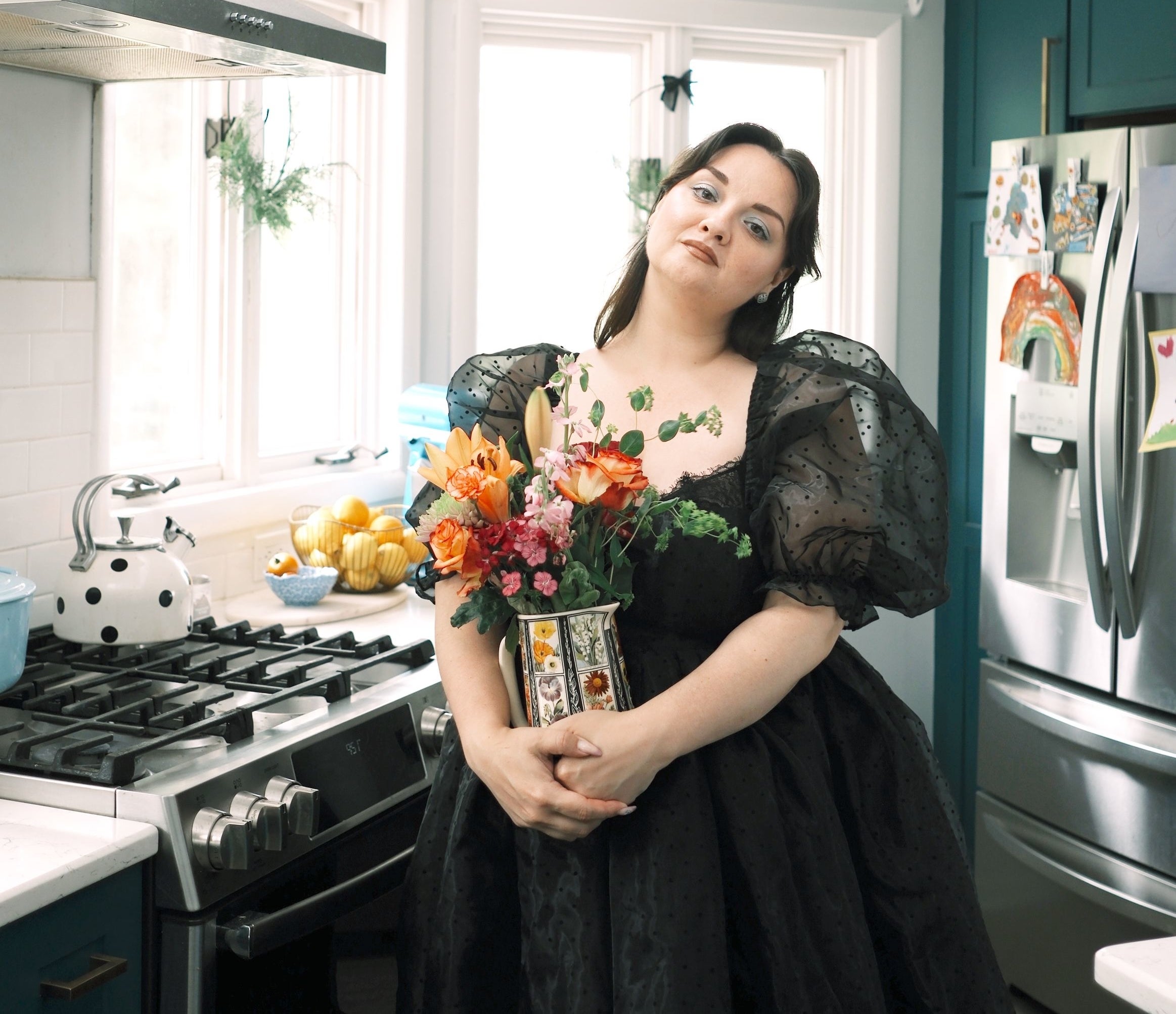 Lynzi stands in her modern kitchen wearing an elegant, puffy-sleeved dress, holding a bouquet of colorful flowers