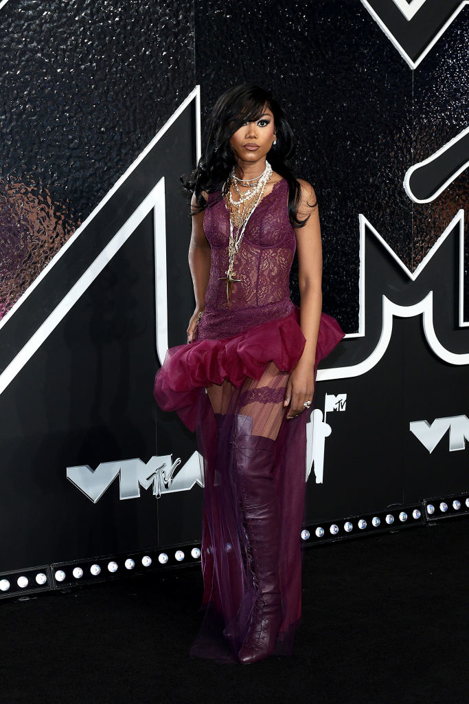 Muni at the VMAs wearing a sheer lace dress with layered tulle, accessorized with multiple necklaces and over-the-knee boots