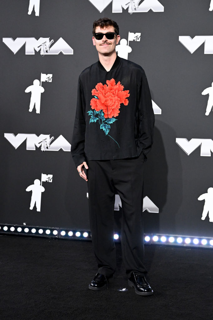 Harry at the VMAs, wearing outfit with a large floral design on the shirt. He wears sunglasses and stands on the red carpet