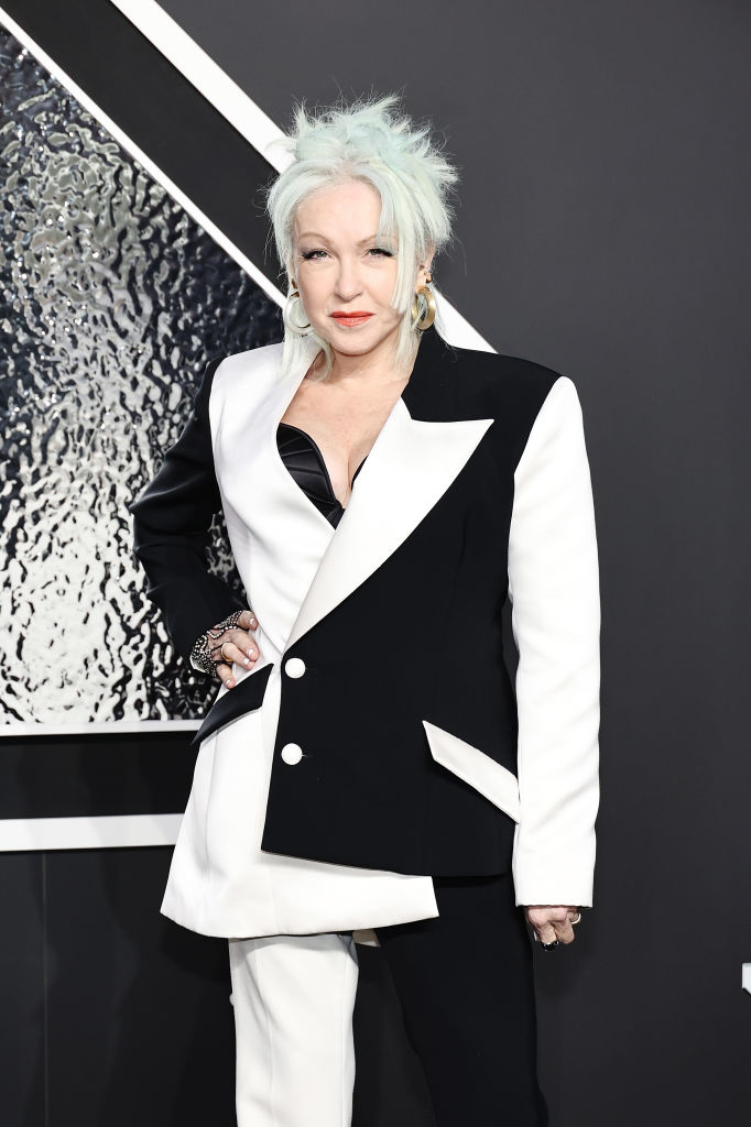 Cyndi Lauper with short hair poses in a stylish asymmetrical blazer suit on the red carpet. She has on large hoop earrings and accessorizes with rings