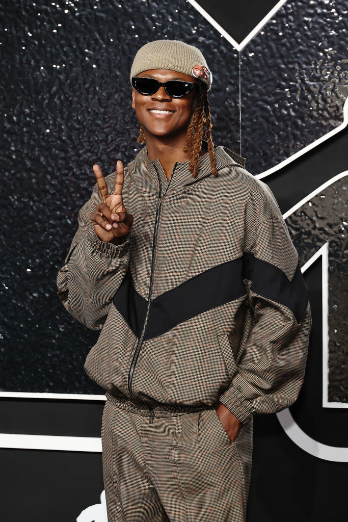 Myles . smiles and flashes a peace sign on the red carpet, wearing a stylish tracksuit, cap, and sunglasses