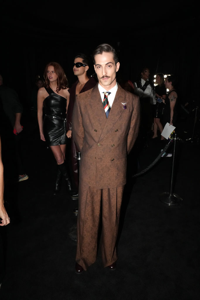 Damiono poses on the red carpet in a double-breasted suit with a patterned tie, while others stand in the background at a celebrity event