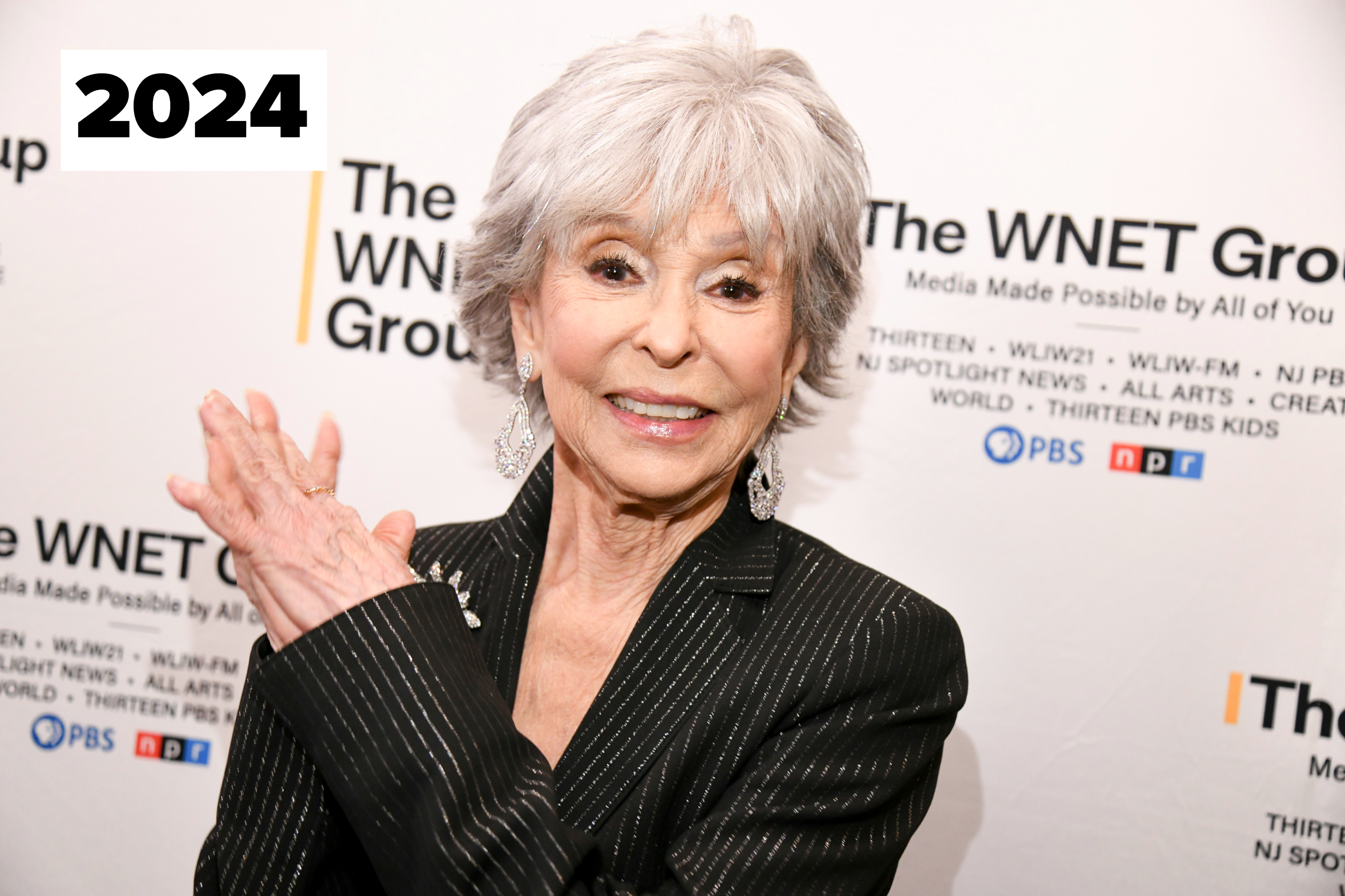 Rita Moreno smiles and poses with her hands together at a WNET Group event. She is wearing a stylish striped suit and dangling earrings