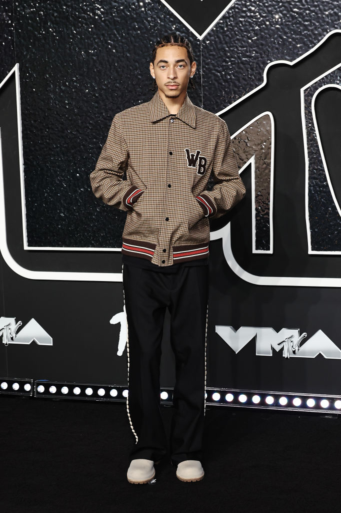 Julez at a VMA event wearing a patterned jacket with "WB" initials, pants, and light-tone shoes
