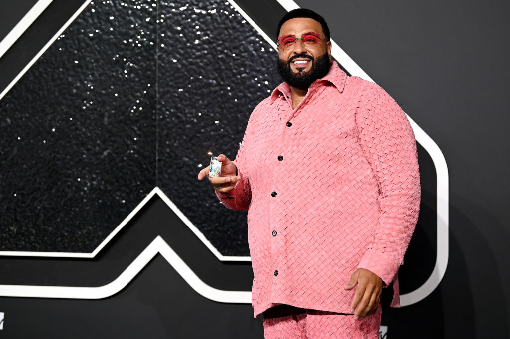 DJ Khaled smiles and poses in an elegant, textured outfit at a celebrity event. He is holding an item in his hand