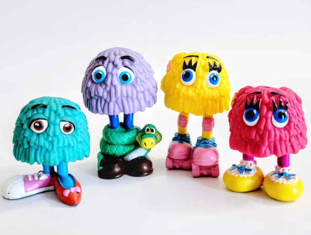 Four colorful fuzzy toy characters with big eyes and various footwear. One holds a green toy snake