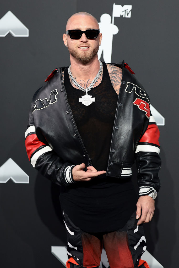 Chet poses on the red carpet wearing a leather jacket with accents, shirt, sunglasses, and multiple silver chains, at an MTV event