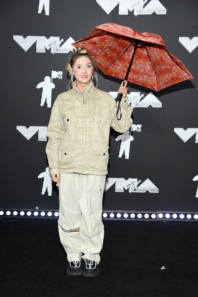 Dora on the red carpet at the MTV VMAs, wearing oversized jacket and pants, holding a patterned umbrella