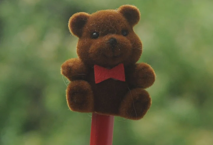 A fuzzy brown teddy bear with a red bow tie is perched on top of a red stick, set against a blurred green background