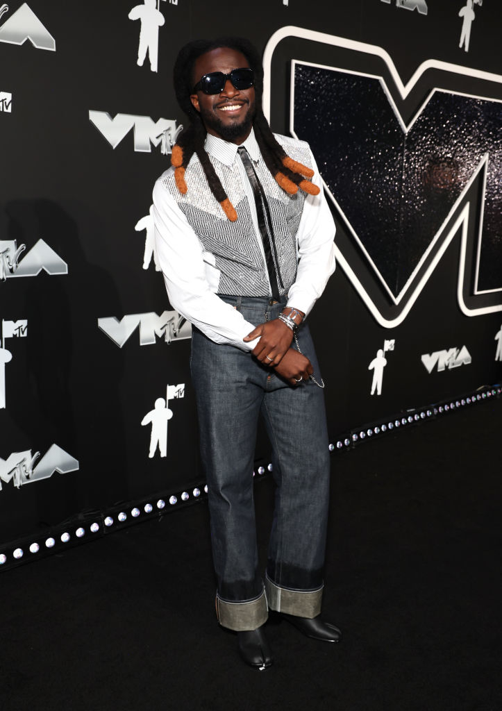 Shaboozey sporting a smile and wearing a striped shirt, wide-legged jeans, and boots on the MTV Video Music Awards red carpet