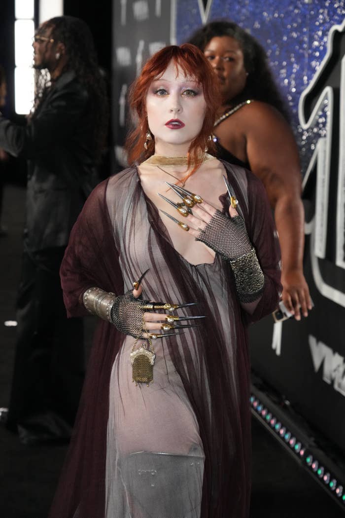 Chappell Roan on the red carpet in a sheer, dark outfit with long sleeves and long, metallic nails. Large dark hoop earrings and layered jewelry complete the look