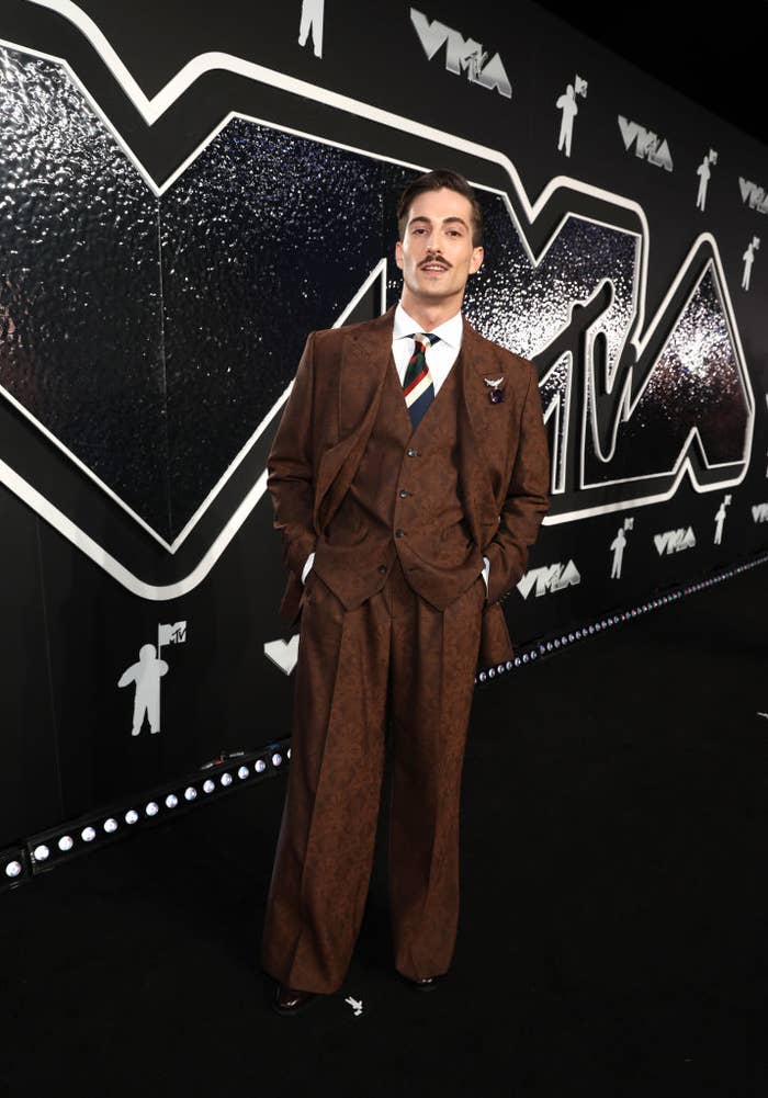 Damiano on the VMAs red carpet in a three-piece suit with a necktie and a brooch on his lapel, posing with hands in pockets