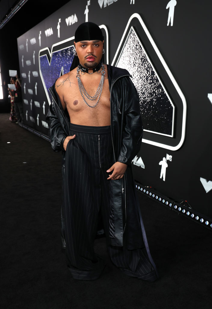 Kandy Muse on a carpet at the VMAs wearing an unbuttoned leather jacket, chains, hat, and pinstripe wide-leg pants