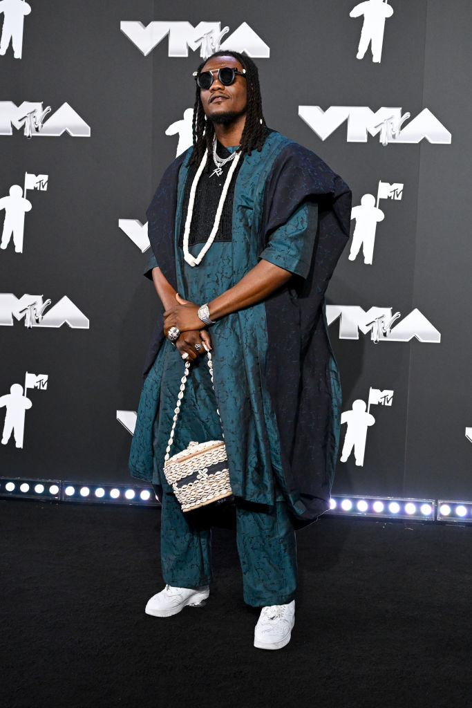 Pheelzl at a celebrity event, wearing oversized attire with a long necklace and holding a woven bag on the red carpet