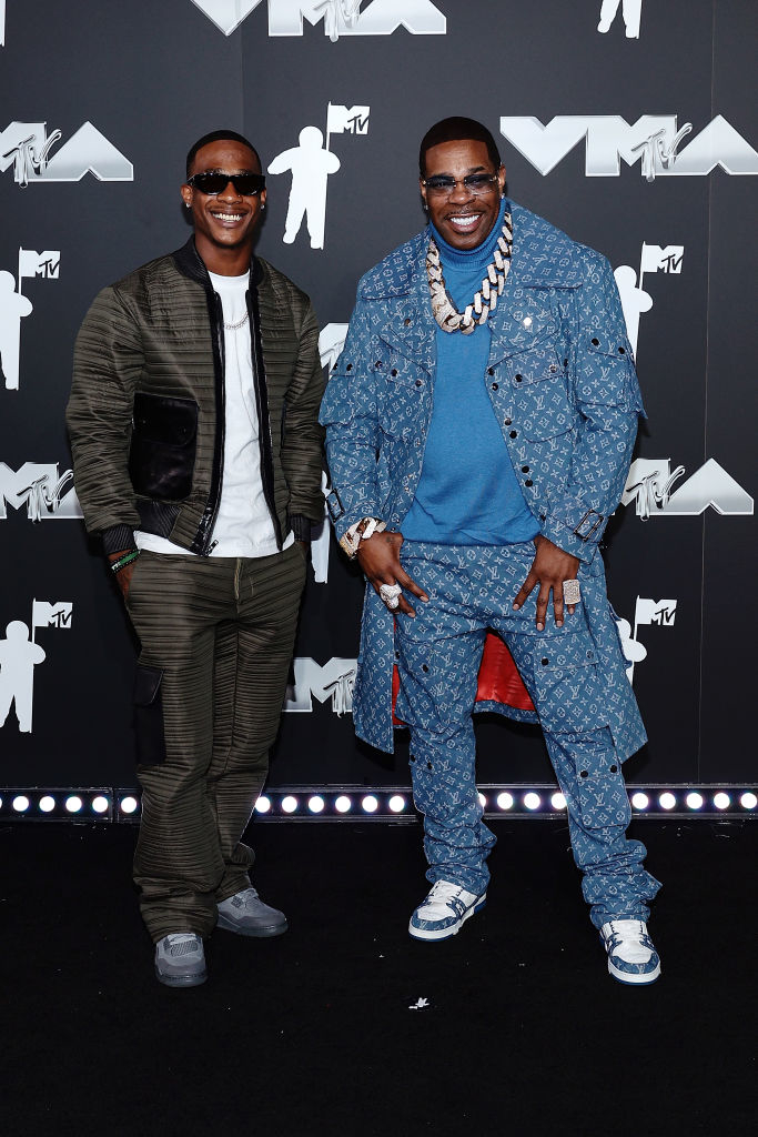 Busta Rhymes and Trilian Wood-Smith are on the MTV VMA red carpet. Busta Rhymes is wearing a denim ensemble with a large chain necklace, and Trillian is in a casual outfit