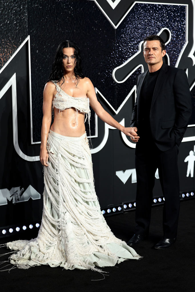 Katy Perry in a draped, two-piece outfit holding hands with Orlando Bloom in a suit at a celebrity event
