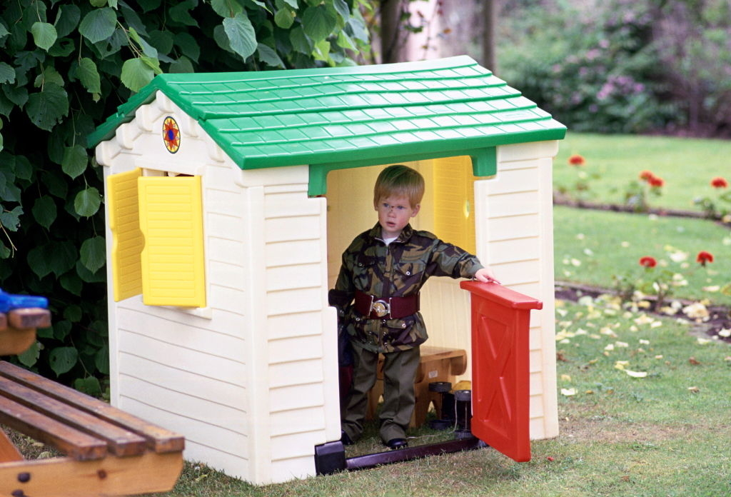 A young boy in a camouflage outfit stands in the doorway of a small plastic playhouse with a green roof and yellow shutters, set in a garden